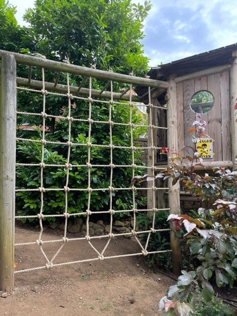 Rope scramble netting on a wooden frame with greenery and a building in the background