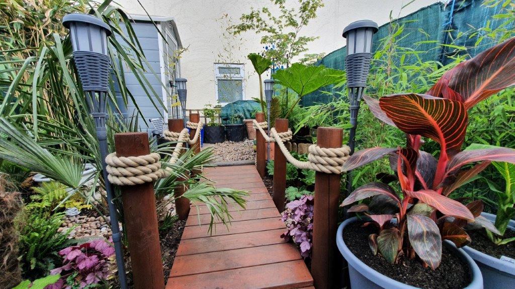 Garden pathway using rope run along posts with wooden planks, plants, and decorative lights.