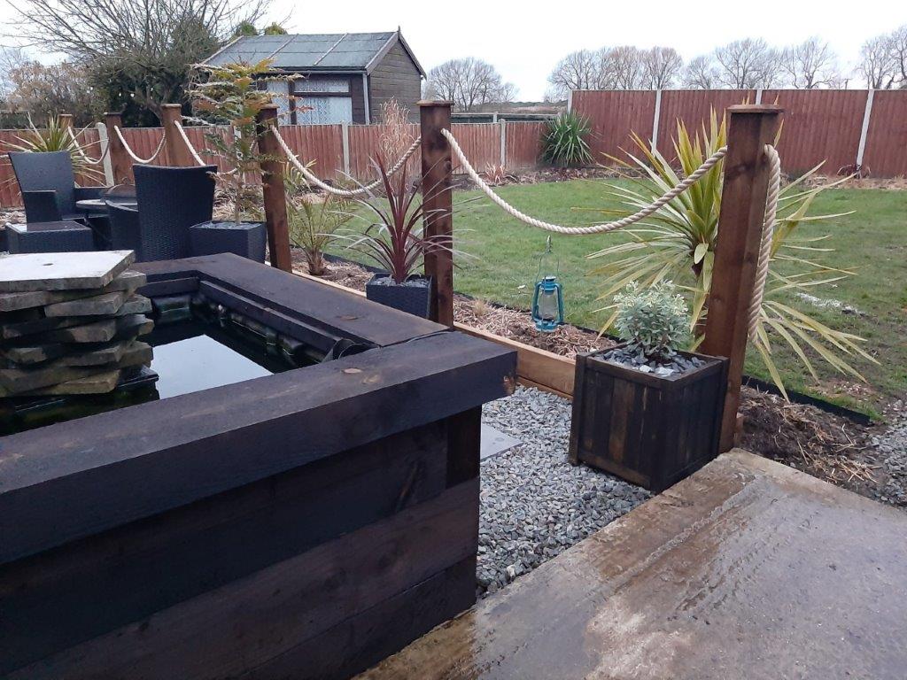 Garden scene showing a rope handrail with a wooden planter, outdoor furniture, and plants.