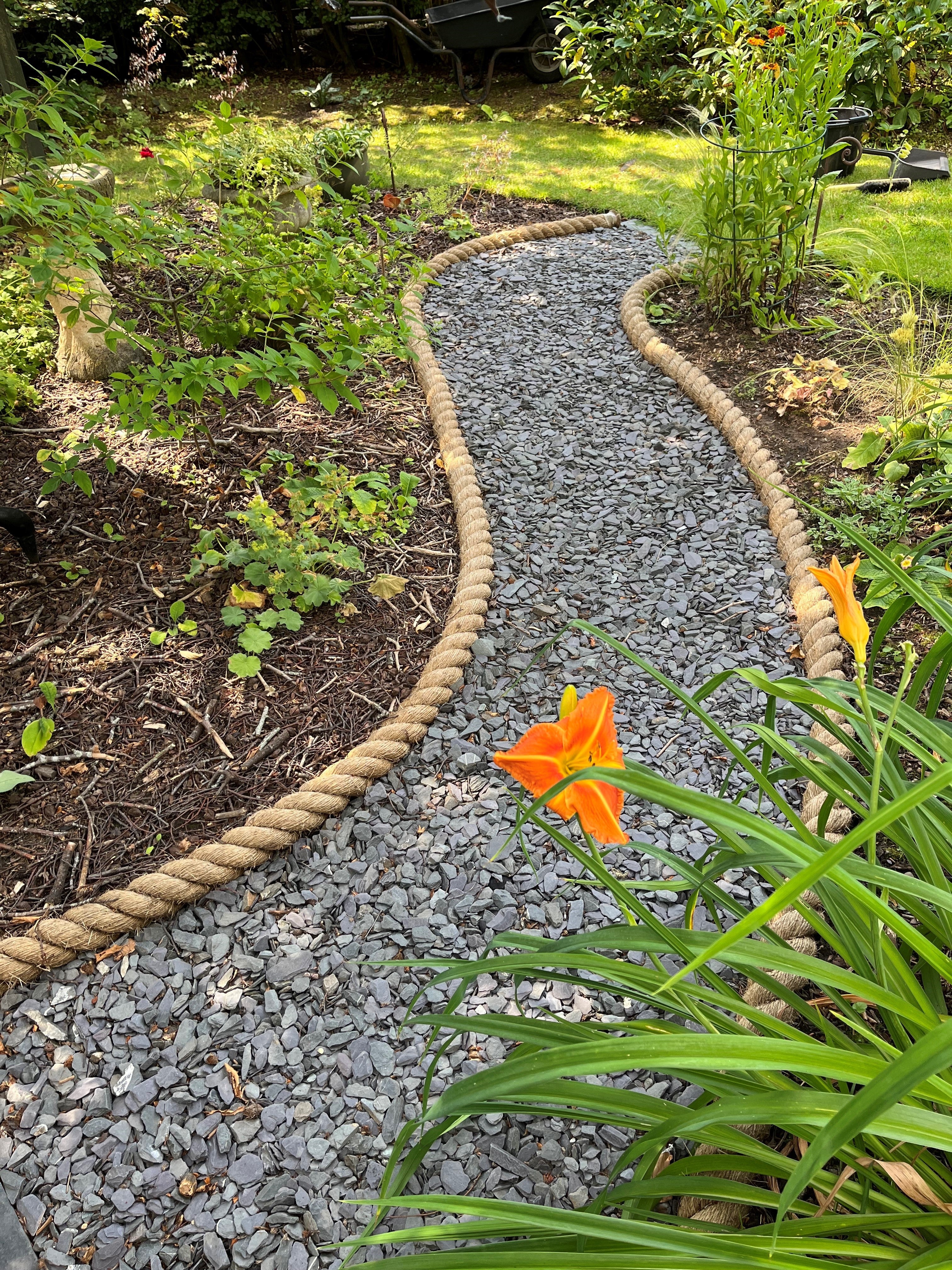 Garden path made of small stones with a manila rope border, surrounded by plants and flowers.